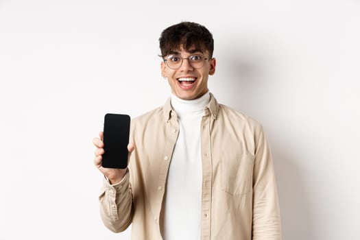 Technology and online shopping concept. Cheerful handsome guy in glasses showing empty smartphone screen and smiling, showing application, standing on white background.