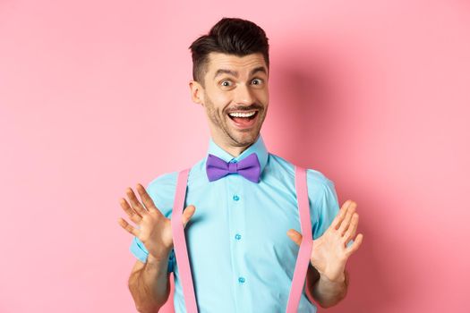 Cheerful guy in bow-tie, showing his suspenders and smiling happy, feeling upbeat, standing excited over pink background.