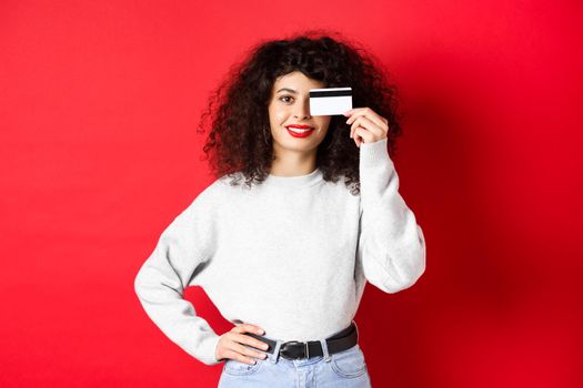 Beautiful female model with curly hair, showing plastic credit card and smiling, standing on red background. Makeup and shopping concept.