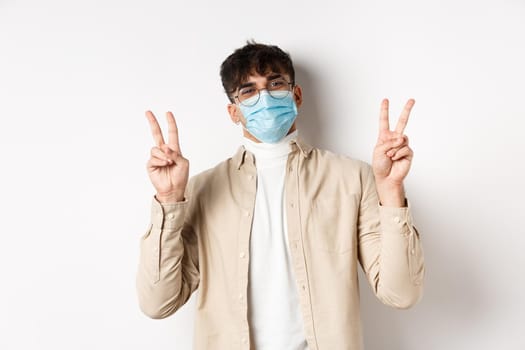 Covid-19, health and real people concept. Cute happy guy in medical mask staying positive, showing peace signs and smiling, standing on white background.