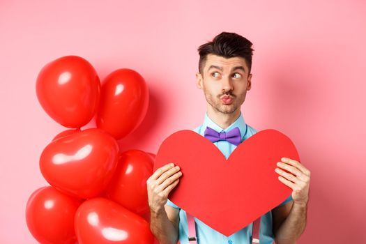 Valentines day concept. Charming young man in bowtie waiting for soulmate with big red heart cutout, looking left and dream of love, pink background.