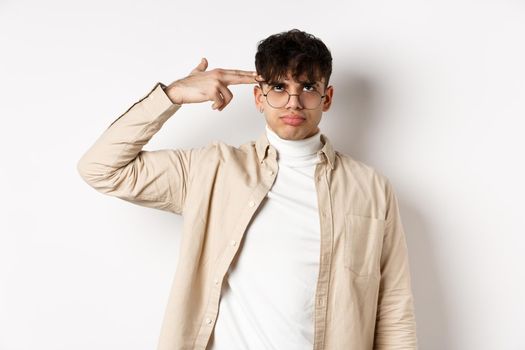 Annoyed young man making finger gun gesture on head, shooting himself from boredom or annoyance, tired of something stupid, standing on white background.