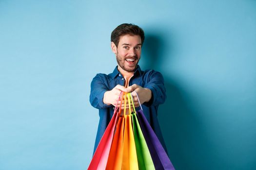 Cheerful young guy stretch out hands with shopping bags and smiling, giving you gifts, standing on blue background.