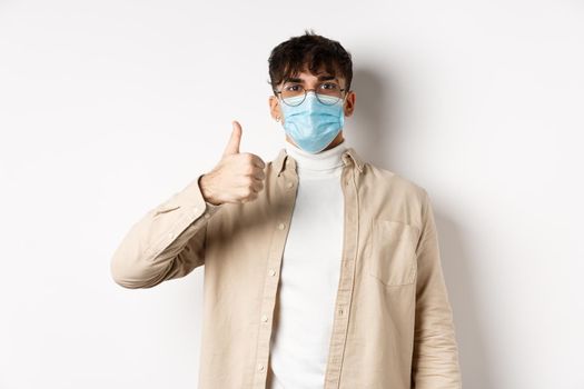 Covid-19, health and real people concept. Satisfied guy in sterile face mask and glasses, showing thumb up in approval, give positive feedback, standing on white background.
