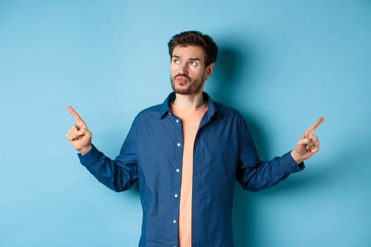 Indecisive man in casual clothes making choice, pointing fingers sideways and looking pensive, standing on blue background.