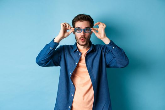 Funny young man trying new sunglasses, looking surprised at camera, standing on blue background. Copy space