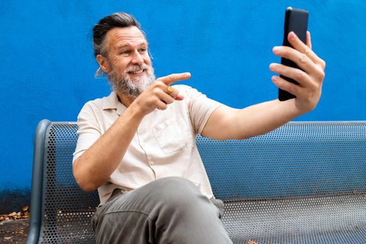 Caucasian mature man with beard sitting on a bench pointing finger to screen during video call. Blue background. Lifestyle concept. Technology concept.