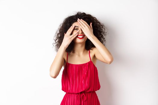 Funny young woman with curly hair, wearing red dress, covering eyes with hands but peeking through fingers and smiling, looking at something interesting, white background.