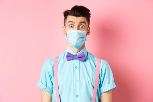 Covid-19, pandemic and health concept. Image of guy looking surprised in medical mask and bow-tie, standing on pink background.