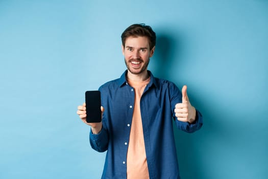 Handsome young man showing thumbs up gesture and empty smartphone screen, recommending app or company, standing on blue background.