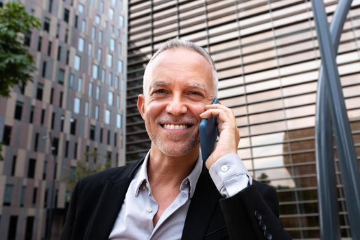 Portrait of smiling caucasian business man talking on cell phone looking at camera next to office buildings. Business and technology concept.
