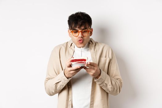 Funny young guy in glasses blowing candle and making wish on birthday cake, standing cheerful against white background.