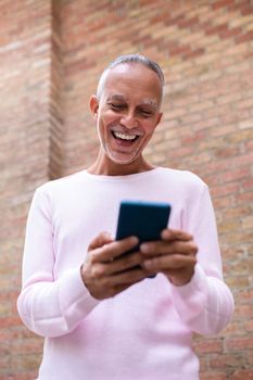 Caucasian adult man using mobile phone outdoors. Browsing through social media. Orange brick wall background. Vertical image. Lifestyle and technology concept.