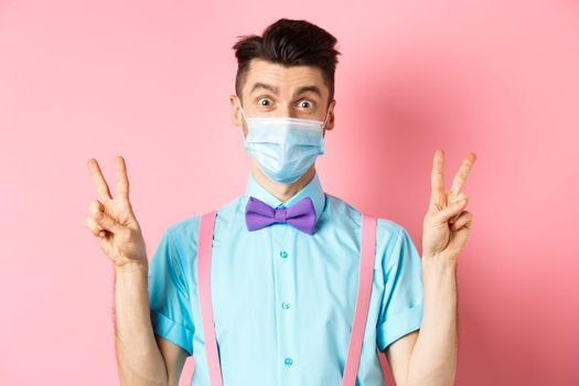 Coronavirus, healthcare and quarantine concept. Funny guy in bow-tie and face mask showing peace, victory signs and looking happy at camera, standing on pink background.