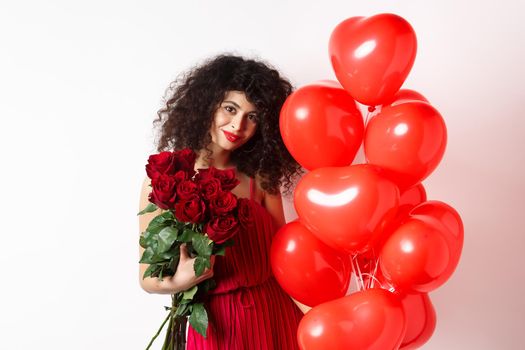 Gorgeous curly haired girlfriend in evening dress, having a date, holding red roses from boyfriend and posing near romantic hearts balloons, white background.