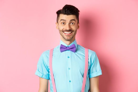 Happy young man with moustache smiling at camera, looking excited and cheerful, standing on pink background in bow-tie and shirt.