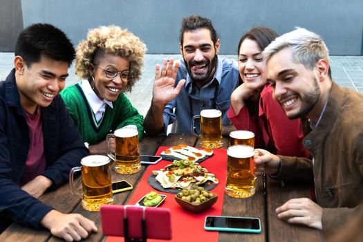 Multiracial group of friends say hello to a friend on a video call using mobile phone. Having beer together in a bar outdoors. Friendship and happiness concept.