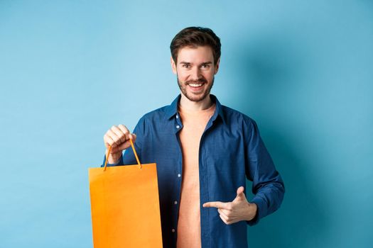 Smiling caucasian man pointing finger at orange shopping bag with bought items, standing on blue background.