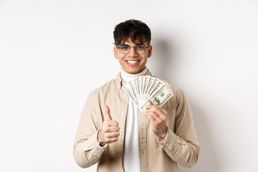 Young guy in glasses showing dollar bills and thumbs up, making money, standing with cash on white background.