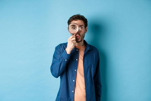 Image of surprised man found something interesting, say wow and look through magnifying glass, standing on blue background.
