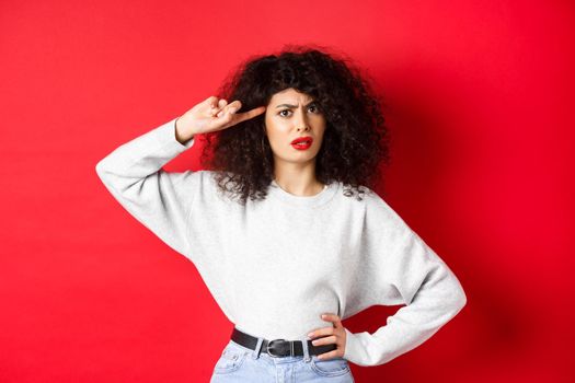 Are you stupid. Annoyed and confused girl with curly hair, pointing at head and scolding person being crazy or strange, standing on red background.