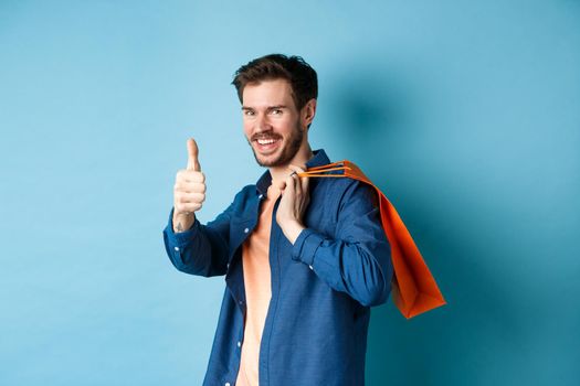 Handsome young guy with shopping bag showing thumbs up and smiling, praising good promotion offer, standing on blue background.