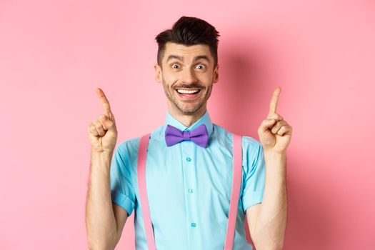 Portrait of cheerful young guy with moustache and bristle pointing fingers up, showing top logo, smiling happy at camera, standing on pink background.