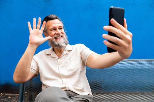 Caucasian mature man with beard sitting on a bench waving hello on video call. Blue background. Lifestyle concept. Technology concept.