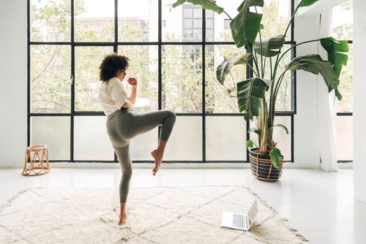 Young multiracial woman kicking leg up doing online fitness training at home living room. Copy space. Active and healthy lifestyle concept.