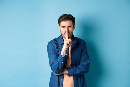 Angry man shushing, press finger to lips and frowning, tell to be quiet, show taboo gesture, standing on blue background.