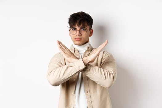 Image of serious young guy in glasses say no, showing cross gesture, make stop sign to disagree or prohibit something, standing on white background.