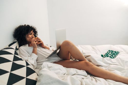 Young mixed race woman lying on bed using laptop and drinking coffee. Copy space. Lifestyle and technology concept.