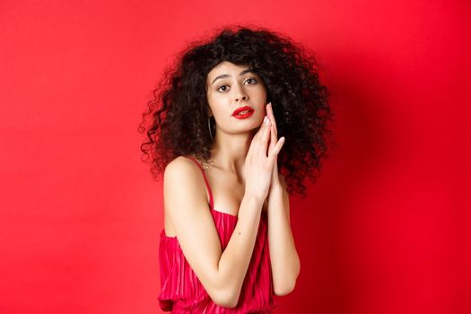 Romantic sensual woman with curly hair, wearing evening dress, posing seductive on red background.