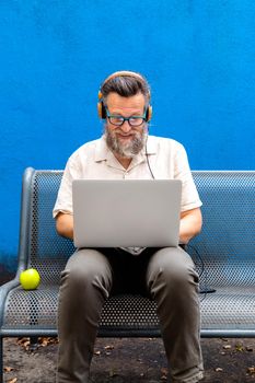 Mature caucasian man wearing headphones using laptop sitting on a bench on blue background. Vertical image. Technology concept.