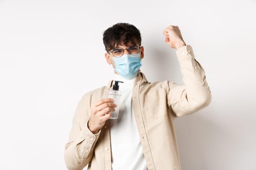 Health, covid and quarantine concept. Happy young man motivated to fight coronavirus, showing hand sanitizer and raising hand up in triumph, standing on white background.