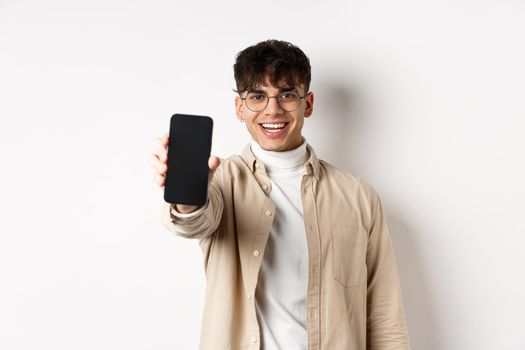 Handsome young man showing empty smartphone screen, standing on white background. Copy space