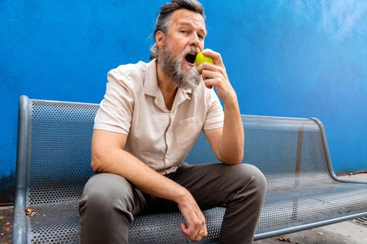 Mature caucasian man with beard eating an apple outdoors looking at camera. blue background. Lifestyle concept.