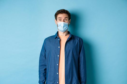 Covid-19 and healthcare concept. Image of handsome modern guy in face mask raising eyebrows in surprise, looking at something in awe, blue background.