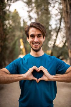 Vertical portrait of happy young caucasian man making heart shape with hands standing in the forest looking at camera. Cardiovascular health concept.