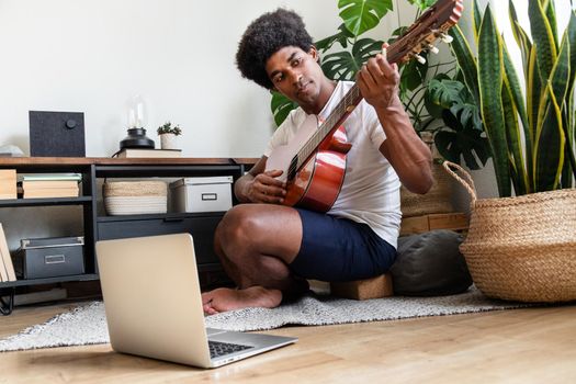 Young african american man learns to play the guitar at home with online course using laptop. Hobbies and leisure activities concept.