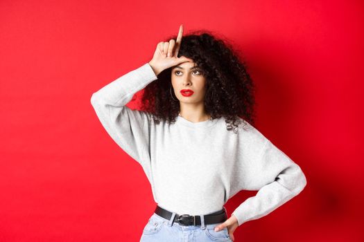 Arrogant woman with curly hair, showing loser sign on forehead and looking aside, mocking someone, standing on red background.