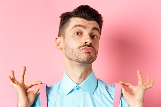 Close up of classy handsome guy adjusting suspenders, pucker lips and raise chin up, looking confident, standing in trendy outfit on pink background.