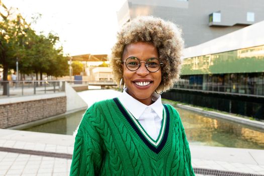 Portrait of smiling young african american woman with blond afro hairstyle, glasses and green jumper in city park. Looking at camera. Copy space. Headshot.