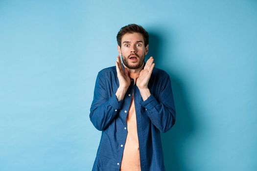 Shocked and scared young man standing in stupor, raising hands up and gasping speechless, standing on blue background.