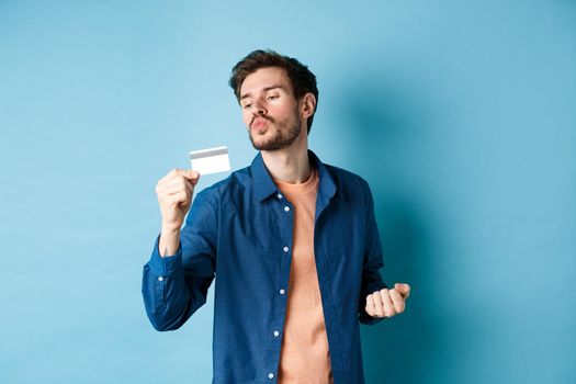 Funny guy kissing his plastic credit card and looking satisfied, shopping with discounts, standing on blue background.