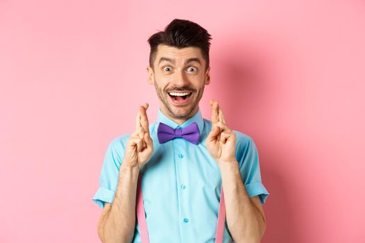 Happy guy making wish, holding fingers crossed for good luck and smiling at camera, waiting for good news, standing over pink background.