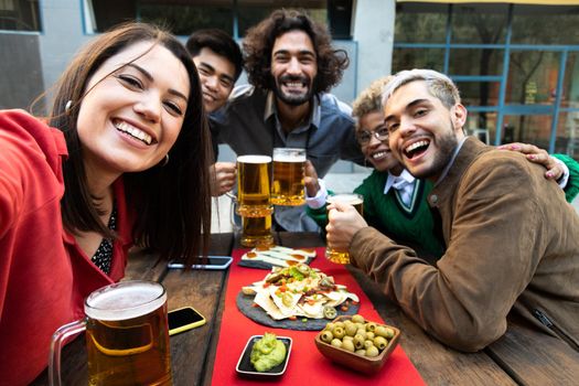 Selfie photo of smiling multiracial friends enjoying some beer and appetizers in a bar outdoors. Friendship and happiness concept. Social media concept.