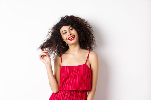 Beautiful woman with curly hair, wearing red dress and lipstick, playing with curl strand and smiling at camera, white background.