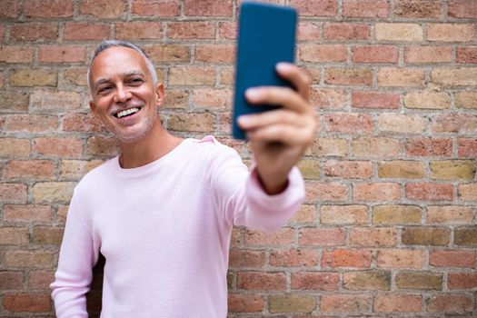 Smiling caucasian adult man taking selfie. Orange brick wall background. Copy space. Technology and lifestyle concept.