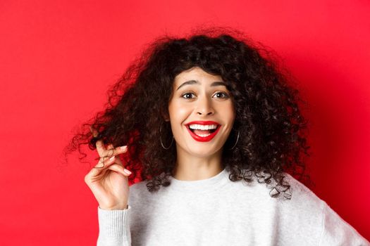 Close-up of happy woman playing with curly hair and laughing at something funny, standing on red background.